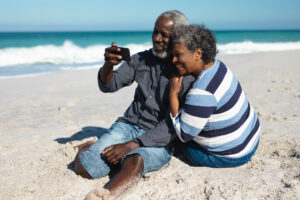 Side view of a senior African American couple reclining on the beach with blue sky and sea in the background, embracing, smiling and using their mobile phone