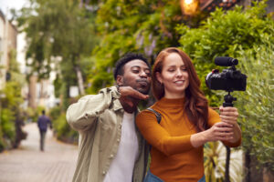 Young Couple Travelling Through City Together Vlogging To Video Camera On Handheld Tripod